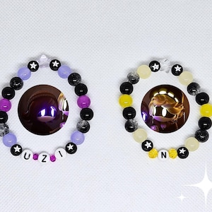 Peut inclure: Deux bracelets de perles. L'un présente des perles violettes, noires et transparentes avec le mot "UZI". L'autre bracelet a des perles jaunes, noires et transparentes avec la lettre "N". Les deux bracelets ont des perles en forme d'étoile.