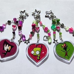 Puede incluir: Tres llaveros en forma de corazón con diseños de personajes de dibujos animados. Cada llavero presenta un corazón de acrílico transparente con un personaje diferente sobre un fondo rojo o verde. Los llaveros tienen detalles de cuentas rosas, moradas y verdes, y dijes en forma de estrella.
