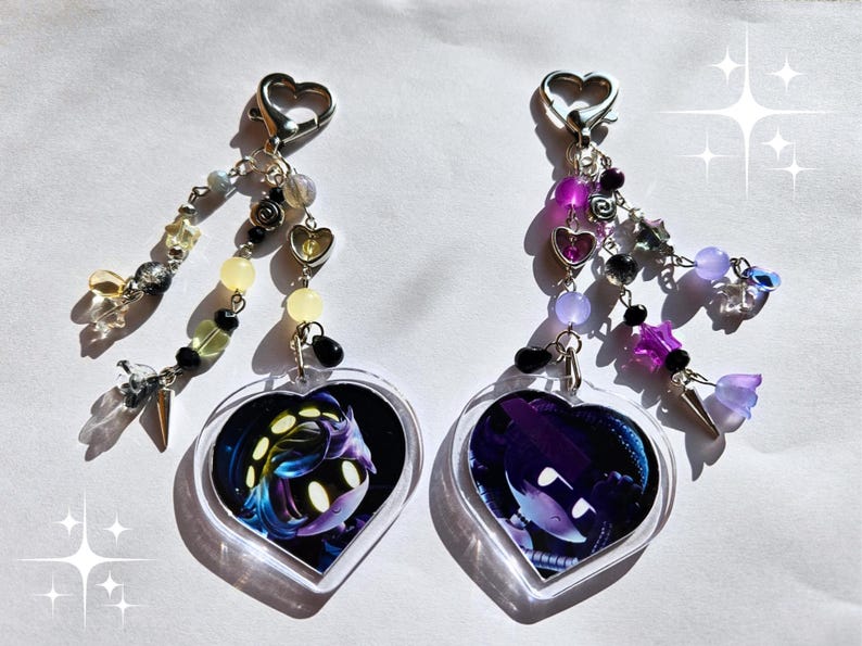 以下が含まれることがあります： キャラクターデザインのハート型キーホルダー2つ。各キーホルダーには、銀色のハート型の留め金と、さまざまな色のビーズチャームが付いています。キャラクターデザインはハートの中心にあります。