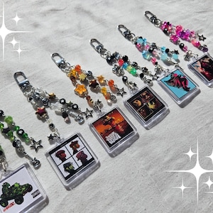 May include: A set of seven keychains featuring various band album covers and a green monster truck design. Each keychain has a silver chain with colourful beads and star charms.
