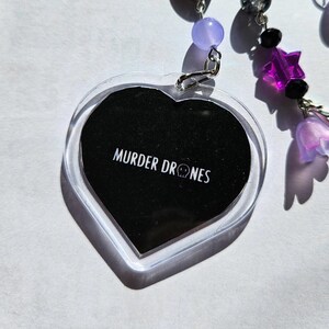 以下が含まれることがあります： 黒い背景に白い文字で「MURDER DRONES」と書かれた、透明なハート型のチャーム。紫と黒のビーズと紫の星型のチャームが付いたチェーンにチャームが取り付けられています。このチャームはファンメイドのアイテムです。