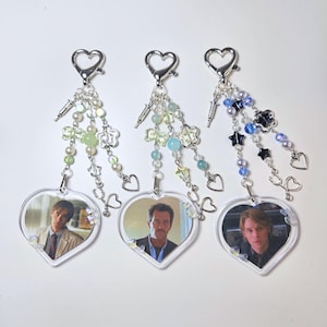 Puede incluir: Tres llaveros en forma de corazón con imágenes de actores. Cada llavero tiene un cierre plateado en forma de corazón, cuentas decorativas y un dije en forma de corazón. Las cuentas son en tonos verdes, azules y blancos.
