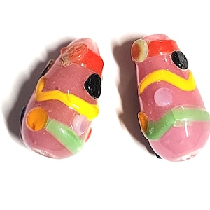 May include: A pair of abstract glass earrings. Each earring is a pink, teardrop shape with colorful accents. The earrings feature red, yellow, green, orange, and black details. The earrings are designed with a unique, artistic style.