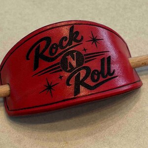 Peut inclure: Une barrette à cheveux en cuir rouge de forme rectangulaire incurvée avec des bords cousus noirs. Le centre présente le texte noir Rock N Roll avec des étoiles. Un bâton en bois clair et lisse est inséré dans deux trous latéraux.