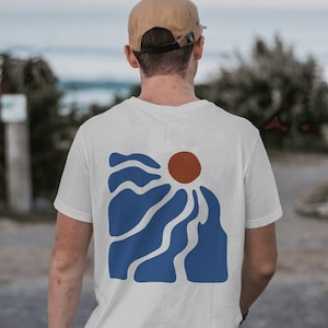 Op de afbeelding: Wit T-shirt met een abstract ontwerp van een zon die opkomt boven een bergketen in blauw en bruin.