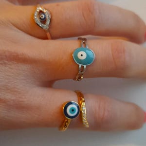 May include: A hand displaying three gold-coloured rings, each with an evil eye motif. The rings have blue and white enamel details. One ring features a teardrop shape with clear stones.