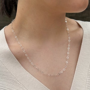 Puede incluir: Un collar de cadena de plata con un delicado dise&ntilde;o de eslabones ovalados. El collar lo lleva una persona con un su&eacute;ter de color claro.