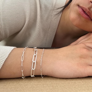 May include: Three silver chain bracelets stacked on a person's wrist. The bracelets are made of different chain styles, including a box chain, a curb chain, and a delicate chain.