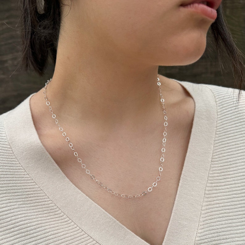 Puede incluir: Un collar de cadena de plata con un delicado dise&ntilde;o de eslabones ovalados. El collar lo lleva una persona con un su&eacute;ter de color claro.