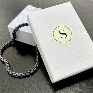 Puede incluir: Un brazalete de cadena negra en una caja blanca con un logotipo que dice "Sincere".
