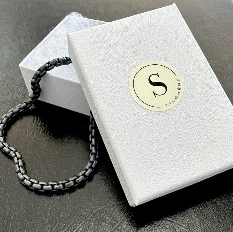 Puede incluir: Un brazalete de cadena negra en una caja blanca con un logotipo que dice "Sincere".