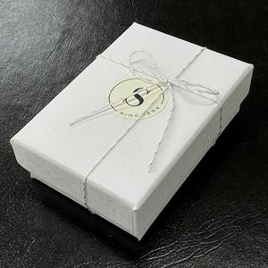 Puede incluir: Una caja de regalo blanca atada con un lazo de cuerda blanca. La caja tiene una pegatina redonda con la letra "S" y la palabra "Sincere" impresa en ella.