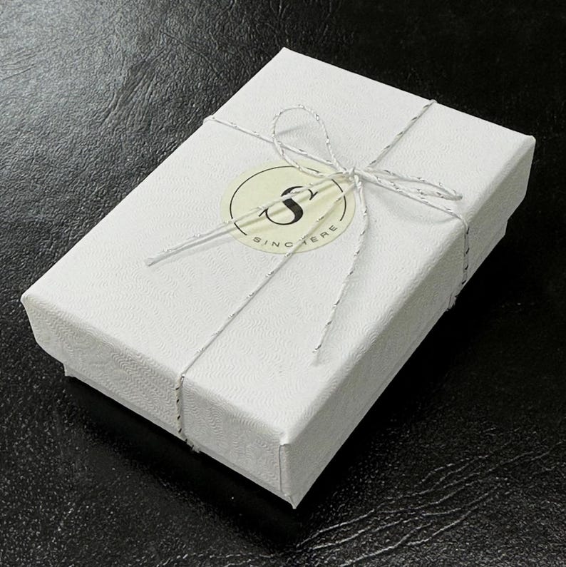 Puede incluir: Una caja de regalo blanca atada con un lazo de cuerda blanca. La caja tiene una pegatina redonda con la letra "S" y la palabra "Sincere" impresa en ella.