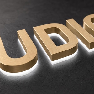 K&ouml;nnte beinhalten: Dreidimensionale Gro&szlig;buchstaben, die das Wort UDIO in einer geb&uuml;rsteten, goldfarbenen Metalloptik bilden. Jeder Buchstabe hat eine dicke Blockschrift mit einem wei&szlig;en LED-Lichtstreifen an der Basis, der einen leuchtenden Halo-Effekt erzeugt.