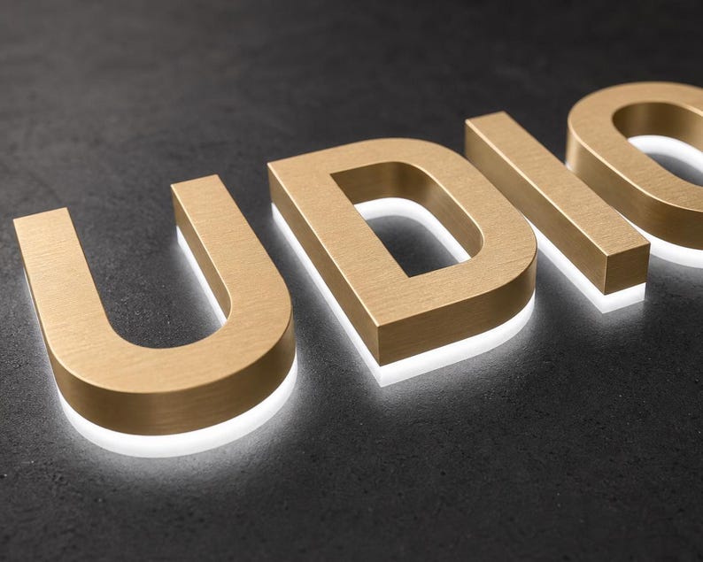 K&ouml;nnte beinhalten: Dreidimensionale Gro&szlig;buchstaben, die das Wort UDIO in einer geb&uuml;rsteten, goldfarbenen Metalloptik bilden. Jeder Buchstabe hat eine dicke Blockschrift mit einem wei&szlig;en LED-Lichtstreifen an der Basis, der einen leuchtenden Halo-Effekt erzeugt.