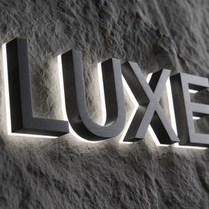 May include: A three-dimensional sign featuring the word LUXE in bold, dark metallic capital letters. Each letter is mounted with a white LED backlight that creates a glowing halo effect around the edges of the characters against the wall.