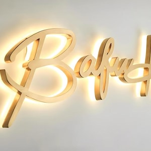 K&ouml;nnte beinhalten: Ein goldfarbenes Metallschild mit dem Wort Bafuh in einer Schreibschrift. Die Buchstaben sind leicht von der Wand abgesetzt montiert, wodurch ein Hintergrundbeleuchtungseffekt mit warmem, leuchtendem Licht entsteht, das die Wand beleuchtet.