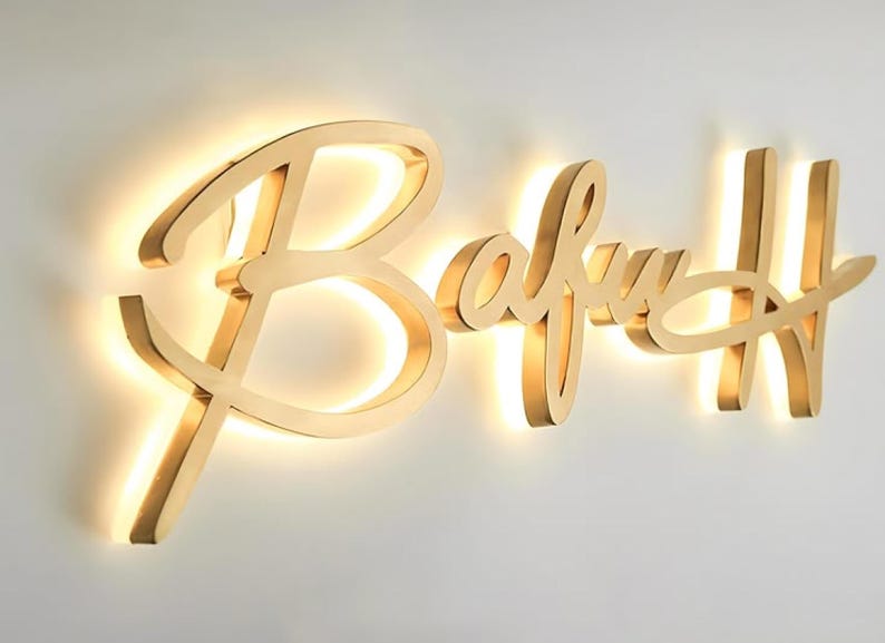 K&ouml;nnte beinhalten: Ein goldfarbenes Metallschild mit dem Wort Bafuh in einer Schreibschrift. Die Buchstaben sind leicht von der Wand abgesetzt montiert, wodurch ein Hintergrundbeleuchtungseffekt mit warmem, leuchtendem Licht entsteht, das die Wand beleuchtet.