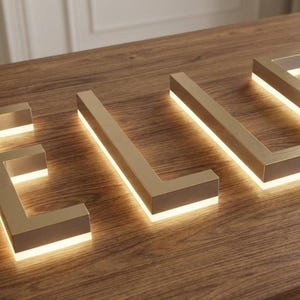 K&ouml;nnte beinhalten: Dreidimensionale Metallbuchstaben, die das Wort ELLE bilden, mit einer geb&uuml;rsteten Goldoberfl&auml;che und integrierter LED-Hintergrundbeleuchtung, die einen sanften Schein unter jedem Zeichen erzeugt und einen Lichteffekt bewirkt.