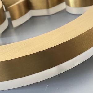 K&ouml;nnte beinhalten: Ein dreidimensionaler, kreisf&ouml;rmiger Schildbuchstabe mit einer geb&uuml;rsteten, goldfarbenen Metalloberfl&auml;che auf der Oberseite und einer massiven wei&szlig;en Acrylbasis. Der Buchstabe hat ein geschwungenes Design mit einer glatten, reflektierenden Textur.