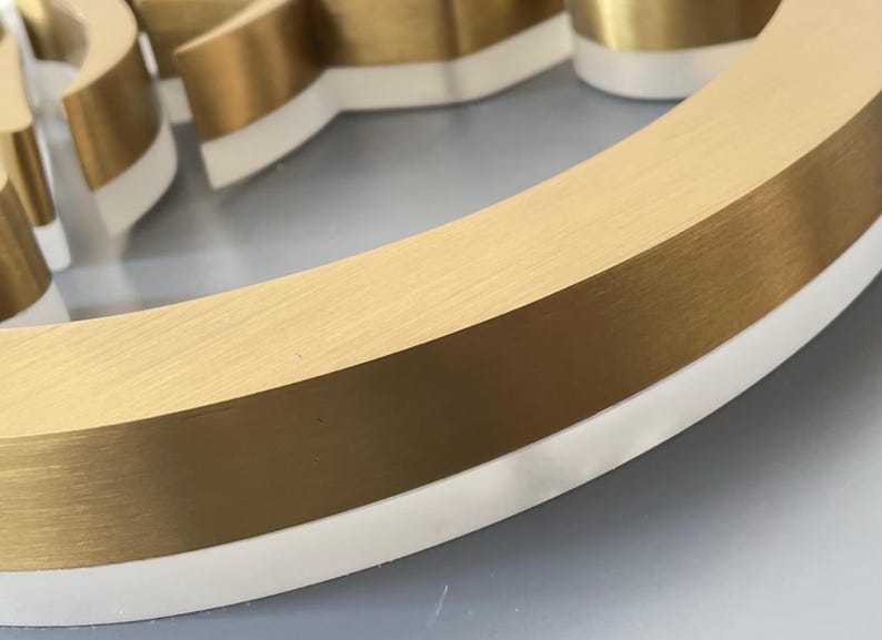 K&ouml;nnte beinhalten: Ein dreidimensionaler, kreisf&ouml;rmiger Schildbuchstabe mit einer geb&uuml;rsteten, goldfarbenen Metalloberfl&auml;che auf der Oberseite und einer massiven wei&szlig;en Acrylbasis. Der Buchstabe hat ein geschwungenes Design mit einer glatten, reflektierenden Textur.