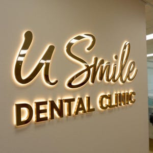 K&ouml;nnte beinhalten: Ein Wandschild mit dem Schriftzug U Smile in goldfarbener Schreibschrift &uuml;ber den Worten DENTAL CLINIC in einer goldfarbenen serifenlosen Schrift. Jeder Buchstabe ist von hinten beleuchtet und erzeugt einen warmen Lichtschein.