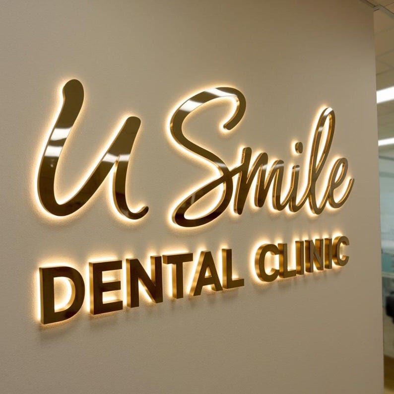 K&ouml;nnte beinhalten: Ein Wandschild mit dem Schriftzug U Smile in goldfarbener Schreibschrift &uuml;ber den Worten DENTAL CLINIC in einer goldfarbenen serifenlosen Schrift. Jeder Buchstabe ist von hinten beleuchtet und erzeugt einen warmen Lichtschein.