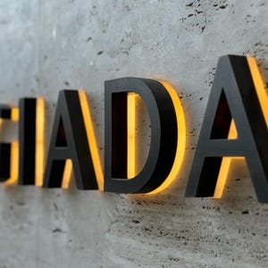 Puede incluir: Letras negras tridimensionales que deletrean "GIADA" con un brillo amarillo cálido que emana de detrás. Las letras están montadas en una pared de hormigón gris texturizado, creando un elemento de diseño moderno.