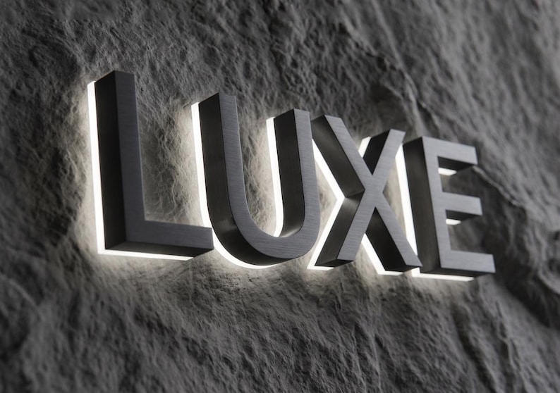 K&ouml;nnte beinhalten: Ein dreidimensionales Schild mit dem Wort LUXE in fetten, dunkelmetallischen Gro&szlig;buchstaben. Jeder Buchstabe ist mit einer wei&szlig;en LED-Hintergrundbeleuchtung versehen, die einen leuchtenden Halo-Effekt um die Kanten erzeugt.