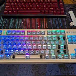 May include: A white mechanical keyboard with a variety of colored keys. The keys are illuminated with blue, green, and red lights. The keyboard has a white frame and a silver metal bar.