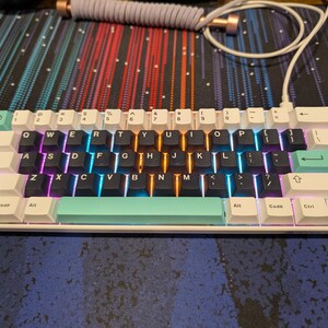 May include: A white mechanical keyboard with black keycaps and a light blue and green accent row. The keyboard has a variety of keys, including function keys, arrow keys, and a number pad. The keyboard is lit up with rainbow colors.