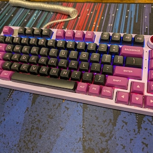 May include: A pink and black mechanical keyboard with a white keycap featuring a red logo. The keyboard has a black space bar and a pink enter key. The keyboard has a pink and black colour scheme.