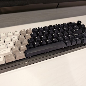 May include: A black and white mechanical keyboard with a clear plastic case. The keyboard has a black and white color scheme with white keys on the left side and black keys on the right side. The keys have white lettering and the keyboard has a black frame.