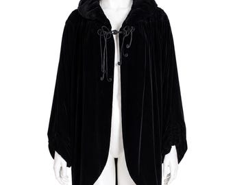 May include: A black velvet cape with a large, rounded collar and a front closure. The cape is open in the front and has wide sleeves. The garment is displayed on a white mannequin.