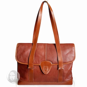 May include: A brown leather tote bag with a flap closure and a single buckle on each side. The bag has two top handles and a brown leather trim.