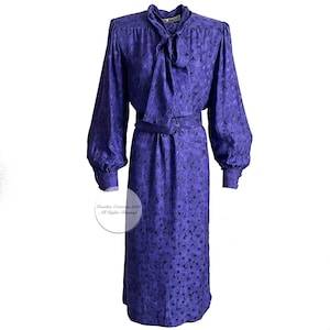 May include: A purple long-sleeved dress with a floral pattern and a tie at the neck. The dress has a belt at the waist.