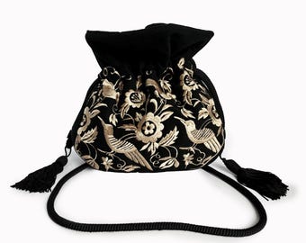 May include: Black drawstring bag with gold floral and bird embroidery. The bag has a black cord strap and two black tassels. The bag's top is gathered with a drawstring closure. The embroidery features detailed floral and bird designs.