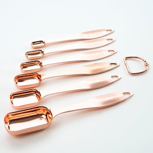 Può includere: Set di sette cucchiaini da misura in oro rosa con manico ad anello. I cucchiaini sono etichettati con misure in frazioni di tazza, tra cui 1/8, 1/4, 1/3, 1/2, 3/4, 1 e 2.