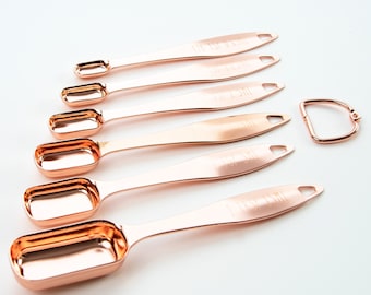 May include: Set of seven rose gold measuring spoons with a ring handle. The spoons are labeled with measurements in fractions of a cup, including 1/8, 1/4, 1/3, 1/2, 3/4, 1, and 2.