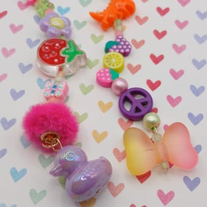 May include: A colorful beaded necklace with various charms, including a strawberry, butterfly, flower, and a peace sign. The necklace features a pink pom-pom, a duck, and a bow. The charms are in shades of pink, purple, orange, and yellow.