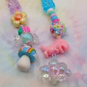 May include: Two colorful beaded necklaces with various charms. One necklace features a flower, strawberry, candy, and mushroom charm. The other necklace has a dinosaur charm and a cluster of clear bubble charms. The beads are in shades of pink, blue, and yellow.