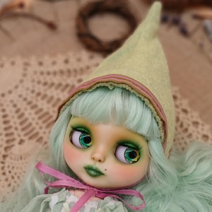 May include: A Blythe doll with mint green hair and large, expressive eyes. The doll wears a light green dress and a pointed hat with pink and green trim. A pink ribbon is tied around the neck.