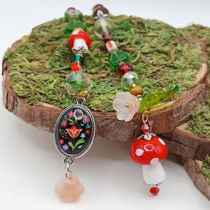 May include: Two beaded charm pendants. One features a red mushroom with white spots, the other an oval pendant with a floral design. The beads are green, red, and clear, with a pink flower and leaf accents.