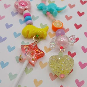 May include: A collection of colorful, whimsical charms and lollipop-style pendants. The charms include a pink flower, a blue dinosaur, a yellow dinosaur, a red star, a candy, an orange slice, and a yellow heart. Each charm is attached to a white stick.