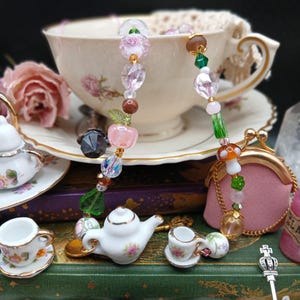 May include: A whimsical beaded charm necklace with a variety of colorful beads, including pink, green, and brown. The necklace is displayed with miniature teacups, a teapot, and a pink coin purse. A vintage teacup and saucer are in the background.