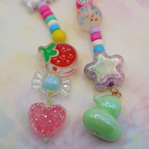 May include: Two beaded charms with colourful beads and charms. One charm features a strawberry, candy, and a pink heart. The other charm has a star and a green duck. The charms are on a light background.