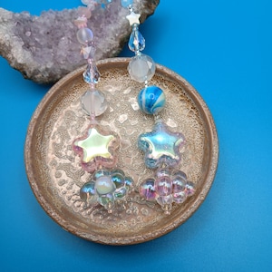 May include: Iridescent star and flower-shaped beaded ornaments with clear and pastel beads. The ornaments are hanging above a decorative, antique-style plate. The background is a solid blue.