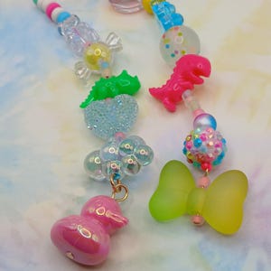 May include: Close-up of colorful beaded jewelry with various charms. The charms include a pink dinosaur, a green heart, a pink duck, a green and yellow butterfly, and other translucent and iridescent beads. The background is a soft, pastel watercolor.