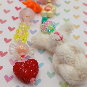 May include: A collection of colorful charms and beads, including a red heart, a white plush bunny, and various candy-shaped pieces. The beads are in shades of pink, yellow, and clear, with a patterned background.