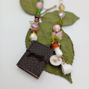 Puede incluir: Un collar caprichoso con un diario en miniatura de cuero marrón atado con un lazo, junto con un dije de hongo, una taza de té y un platillo decorado con flores. El collar incluye cuentas rosas, verdes y marrones, y un dije de mariposa rosa.